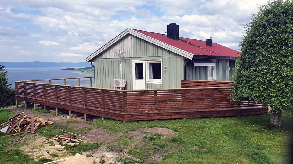 A grey-green house with a red roof and a new terrace with a slatted rail, the water behind